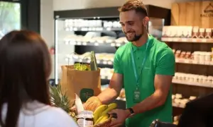 Sprzedawca w zielonej koszulce podaje klientce koszyk z zakupami w sklepie spożywczym.