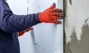 Ręce w pomarańczowych rękawicach przytwierdzają płyty styropianowe do ściany, wykonując ocieplenie stropu styropianem.