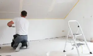 Mann kniet und spachtelt Deckenfugen, um Rigipsplatten zu kleben. Eine Leiter und ein Eimer stehen bereit.
