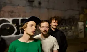 Trzech mężczyzn na tle graffiti. Jeden w zielonej bluzie i czapce, drugi w białej koszulce, trzeci w czarnej kurtce z wąsami. Ich łona niekonieczny krupa.