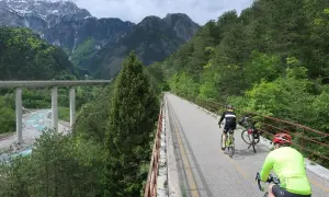 Rowerzyści przemierzają malowniczą trasę **Alpe Adria** w otoczeniu gór i lasów. Widoczny most nad rzeką dodaje krajobrazowi majestatu.