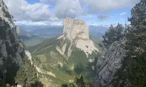 Majestätischer Mont Aiguille ragt aus grünen Tälern empor, umgeben von Felsen und Kiefern unter einem dramatischen Wolkenhimmel.