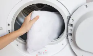 A hand places a white pillow into a washing machine, demonstrating how to wash pillows in the washing machine for a fresh, clean sleep.