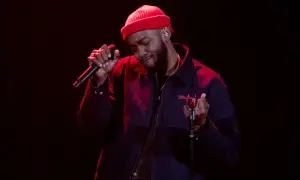 Ant Clemons, wearing a red beanie and dark jacket, sings passionately into a microphone on a dimly lit stage.