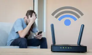 Zmartwiony mężczyzna siedzi na kanapie, trzymając telefon. Obok router Wi-Fi z ikoną sygnału, sugerującą słabą siłę sygnału wifi.