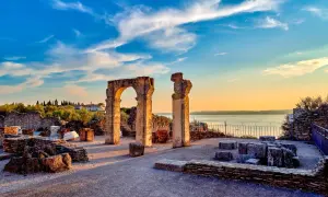 Resti archeologici delle Grotte di Catullo con un arco in pietra e colonne, sullo sfondo il lago e un cielo azzurro al tramonto.
