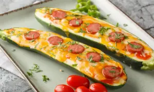 Dwie połówki cukinii faszerowanej warzywami i mozzarellą, zapieczone z plasterkami kiełbasy i posypane ziołami.