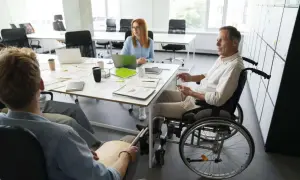 Spotkanie zespołu, gdzie mężczyzna na wózku inwalidzkim, mimo znacznego stopnia niepełnosprawności, aktywnie uczestniczy. Przywileje w pracy to dostępność i równość.