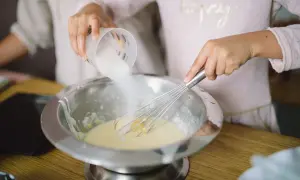 Préparation de la pâte à crêpes : du sucre est versé dans un saladier où une main fouette la préparation.