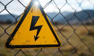 Gelbes Warnschild mit Blitzsymbol auf einem Maschendrahtzaun, das auf die Gefahren des elektrischen Stroms hinweist.