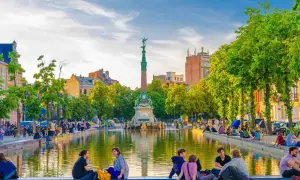 Des gens profitent d'une journée ensoleillée au bord d'un étang, un monument imposant se dresse en arrière-plan. Une belle scène pour visiter Bruxelles.