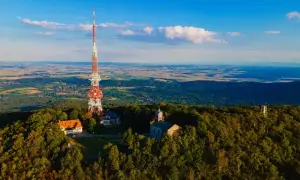 Wieża RTV na górze Ślęża góruje nad lasami i krajobrazem. Obok niej budynki i kościółek.