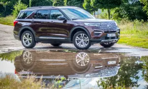 Ciemnobordowy Ford Explorer, jeden z popularnych ford suv modele, odbija się w kałuży po deszczu, otoczony zielenią.
