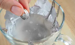 Dłoń trzyma srebrny pierścionek nad szklaną miską z wodą i folią aluminiową, obok wisi srebrny łańcuszek.