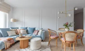 Miękka, błękitna sofa i pudroworóżowe akcenty tworzą przytulną atmosferę. Ciepłe kolory do salonu podkreślają drewniane meble i jasny dywan.