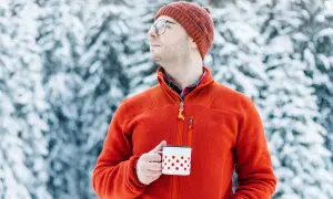 Mężczyzna w pomarańczowym polarze i czapce trzyma kubek w śnieżnym lesie. Polar jest wykonany z miękkiego, ciepłego materiału.