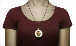 Okrągły medalion z bukietem maków na brązowym łańcuszku, noszony na bordowej bluzce.