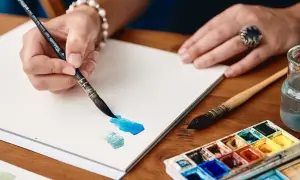 Mano che dipinge con acquerelli su un sottilissimo foglio per acquerelli e litografie. Tavolozza di colori e pennelli vicini.