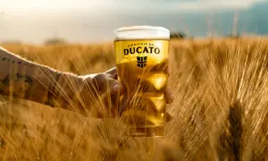 Un bicchiere di birra dorata, con schiuma bianca, tenuto in mano tra spighe di grano mature. Un assaggio di birra al cioccolato, con il logo Ducato.