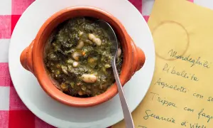 Zuppa di cavolo nero e fagioli in ciotola di terracotta, servita su un piatto bianco con tovaglia a quadri rossi. Accanto, un foglio giallo con un elenco di piatti.