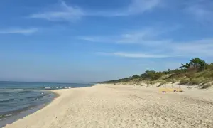 Słoneczny dzień nad zalewem kamieńskim. Szeroka, piaszczysta plaża z łagodnymi falami i zielonymi wydmami w tle. Kilka osób odpoczywa na piasku.