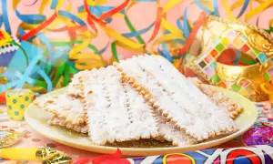 Bugie ricetta originale, dolci fritti cosparsi di zucchero a velo, su un piatto con coriandoli e maschere di Carnevale.