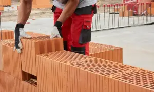 Budowlaniec w czerwonych spodniach i rękawicach układa pustaki. Cegły tworzą ścianę, a w tle widać materiały budowlane i zbrojenie.