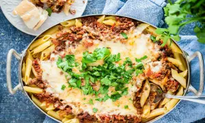 En stor ugnsform med gyllene penne, täckt med köttfärssås och smält ost. En portion pasta al forno är upplagd på en tallrik bredvid.