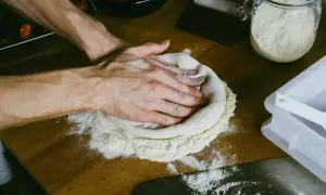 Dłonie formują ciasto na pizzę na drewnianym blacie. W tle słoik z mąką i sos pomidorowy. Zastanawiasz się, ile ma rosnąć ciasto na pizzę?