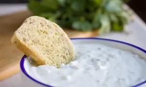 Ein Stück Brot wird in eine Schüssel mit cremigem, selbstgemachtem Zaziki getunkt. Frische Kräuter im Hintergrund.