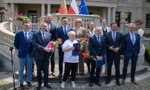 Grupa osób w garniturach i strojach formalnych stoi przed budynkiem z flagami.