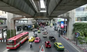 Tętniący życiem transport w Bangkoku: czerwony autobus, żółte i różowe taksówki, samochody i motocykle na ruchliwej ulicy pod betonową estakadą.