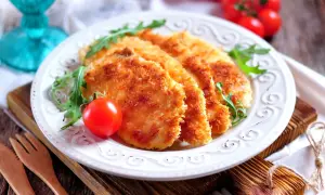 Delizioso pollo impanato al forno, croccante e dorato, servito con pomodorini e rucola su un piatto bianco.