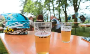 Bicchiere di birra con logo White Dog Brewery, accanto a un casco da moto blu, su un tavolo all'aperto.