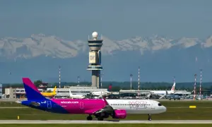 Różowy samolot Wizz Air na płycie lotniska, z wieżą kontroli lotów i ośnieżonymi górami w tle. Jak wydrukować bilety Wizzair?