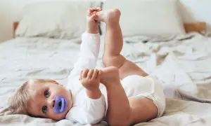 En 11 månaders bebis med napp ligger på rygg på en säng och leker med sina fötter.