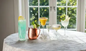 Fem vackra, enkla drinkar står uppradade på ett dukat bord. En blå, en gul, en klar, en med skum och en ljusgrön, alla med garnityr.