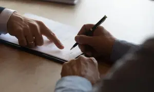 Podpisanie umowy, gdzie jeden mężczyzna wskazuje palcem na dokument, a drugi składa podpis. To ważny krok, by zabezpieczyć zadatek na mieszkanie.