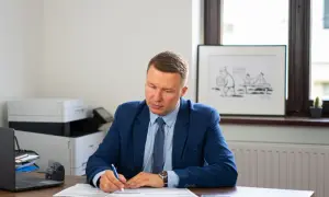 Mężczyzna w garniturze siedzi przy biurku i pisze na dokumentach.