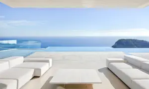 Terrasse moderne avec canapés blancs et table basse, bordée par une piscine à débordement et la mer bleue. Le **béton ciré autour de la piscine** crée une ambiance épurée et luxueuse.