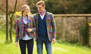 Ein Paar in farbenfrohen Tweed-Jacken und Westen spaziert durch einen sonnigen Park. Was ist Tweed? Ein robuster, traditioneller Stoff.