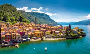 Village coloré au bord des lacs italiens, avec des montagnes verdoyantes en arrière-plan et un voilier sur l'eau turquoise.