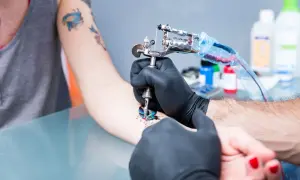 Artysta w czarnych rękawiczkach wykonuje tatuaż motyla na przedramieniu. W tle widać inne tatuaże i butelki z płynami, sugerujące proces gojenie tatuażu na sucho.
