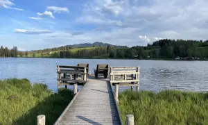 Holzsteg mit Sitzgelegenheiten am bayerischen See, umgeben von grünen Wiesen und bewaldeten Hügeln unter blauem Himmel.