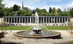 Ein Brunnen sprudelt vor einem modernen Gebäude mit Säulen und Glasfronten. Im Hintergrund ein Kirchturm und Bäume. Ein idyllischer tschechischer Kurort.