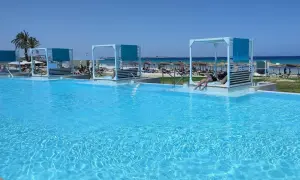 Basen z turkusową wodą, leżaki z baldachimami i plaża w tle.