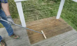 Mann reinigt mit einem Besen die Holzterrasse. Die eine Hälfte ist schon nass und dunkler, die andere noch trocken.
