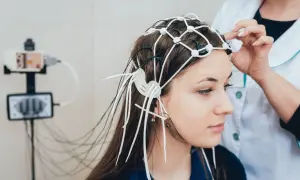 Młoda kobieta z czepkiem EEG na głowie, przygotowywana do badania. W tle aparat do zapisu aktywności mózgu.