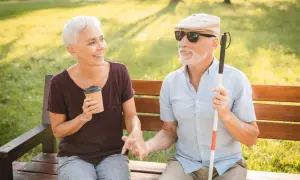Coppia anziana al parco, lui con bastone bianco e occhiali da sole, lei con caffè. Parlano serenamente, forse di come affrontare l'otticopatia.