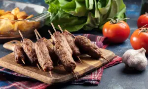 Aksamitne cevapcici z grilla, podane na desce z dodatkami: pieczone ziemniaki, pomidory i czosnek.